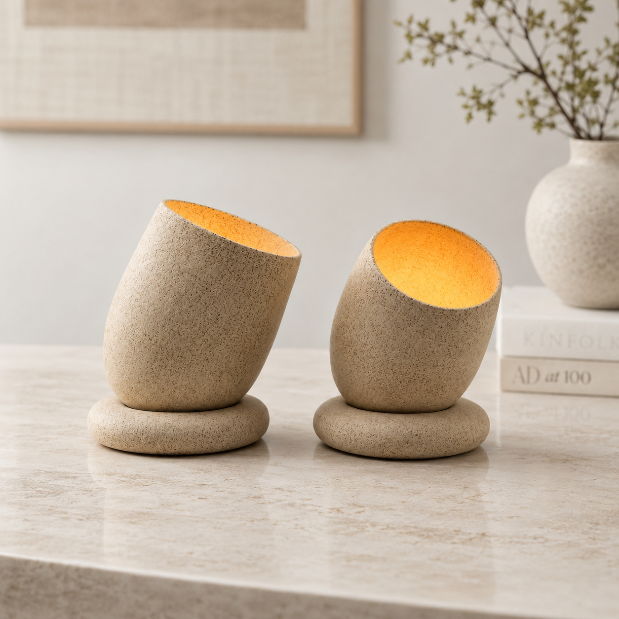 Set of two tilted ceramic tealight holders in sandy cream with warm amber glow on a marble surface