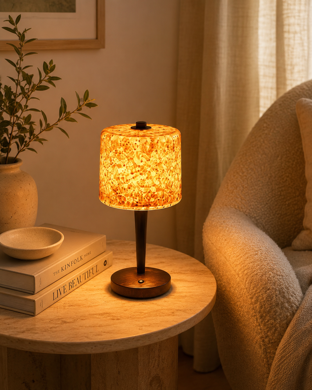 Tortoiseshell glass table lamp glowing warm amber on a marble side table with books and ceramic bowl next to a linen armchair