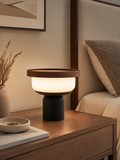 Walnut tray table lamp with opal glass globe glowing on a dark wood bedside table with dried pampas grass and ceramic dish