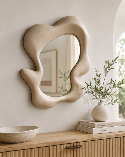 Abstract wave wall mirror with sandy cream organic frame above a ribbed oak sideboard with ceramic vase and books