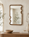 Wavy walnut wood frame wall mirror above a ribbed oak console with wooden bowl and white ceramic vase with dried stems