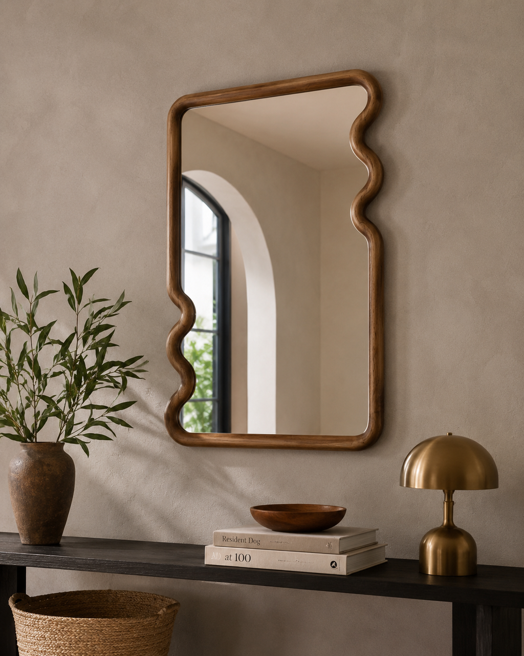 Wavy wood frame mirror on a warm beige wall above a dark console with terracotta vase, wooden bowl and brass mushroom lamp