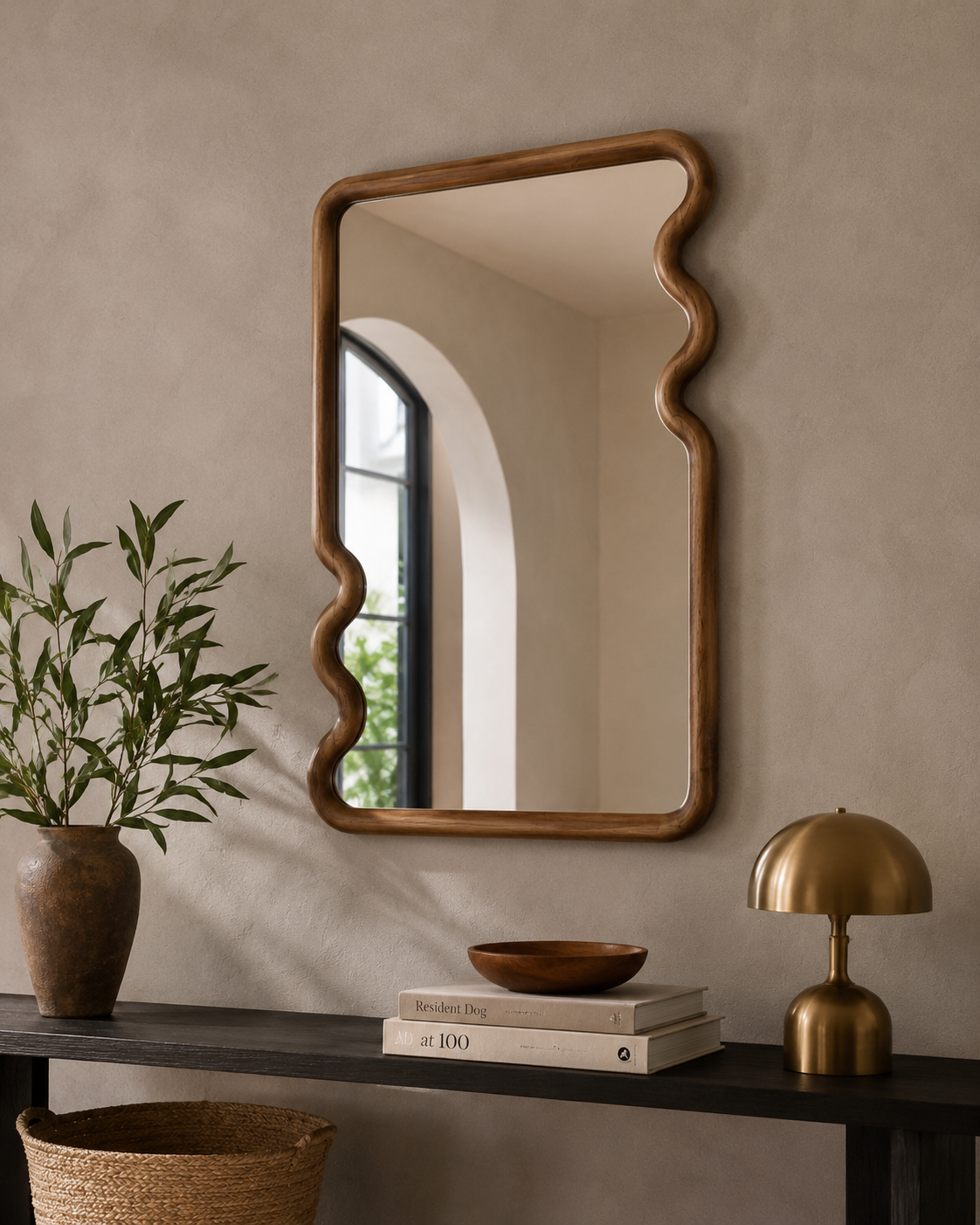 Wavy wood frame mirror on a warm beige wall above a dark console with terracotta vase, wooden bowl and brass mushroom lamp